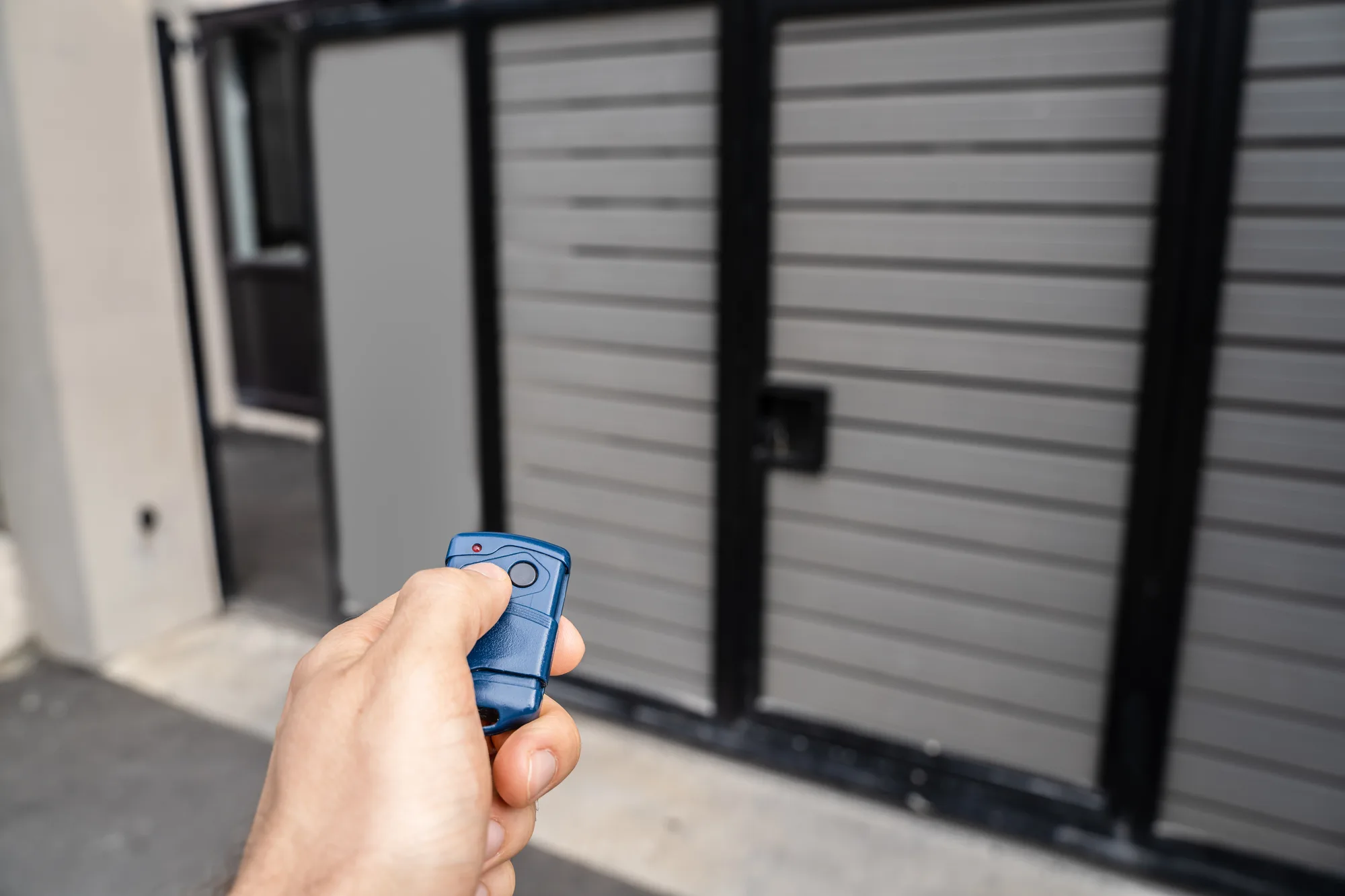 Hand holding a gate remote control in front of a grey automatic gate